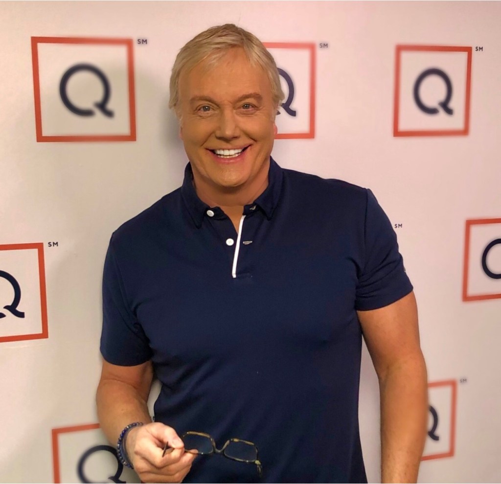 Rick Domeier smiles at a QVC event and holds a pair of glasses in his hand. Behind him is a wall with QVC's logo.