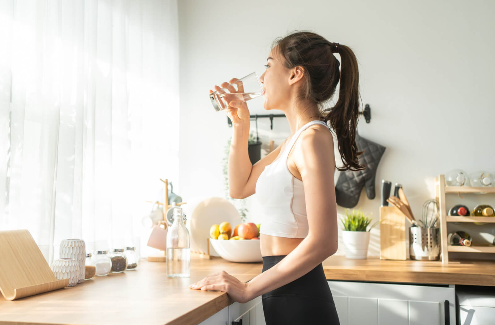 A woman in her sportswear drinking a glass of water in a brightly lit kitchen.