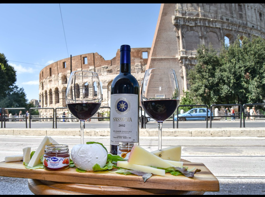 A bottle of wine and cheese on a table with a building in the background

Description automatically generated