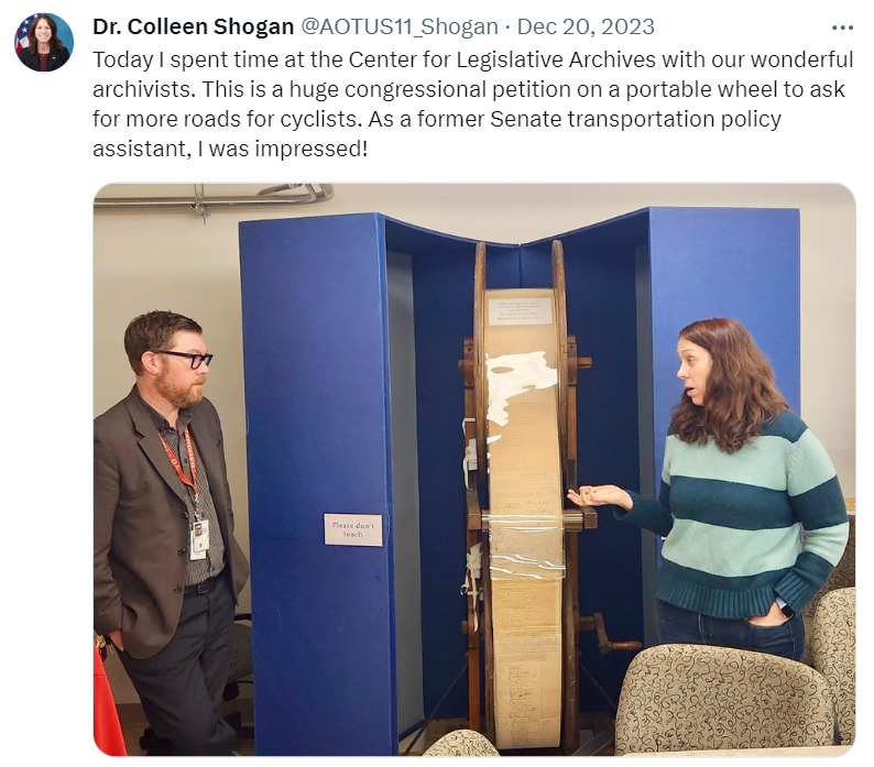A screenshot of an X post from Dr. Colleen Shogan that says, "Today I spent time at the Center for Legislative Archives with our wonderful archivists. This is a huge congressional petition on a portable wheel to ask for more roads for cyclists. As a former Senate transportation policy assistant, I was impressed!" Below is in image of Dr. Shogan next to the wheel display.