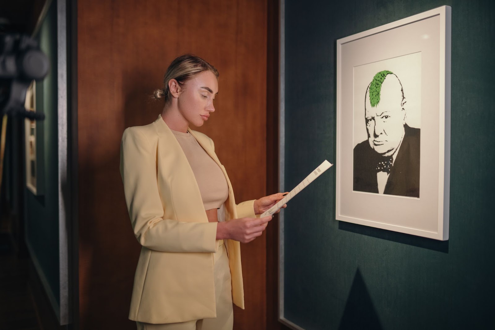 Gallery, Irina Garasymiv, founder of 10101.art, studies a brochure next to Banksy's 'Turf War,' a black and white portrait of Winston Churchill with a green mohawk.