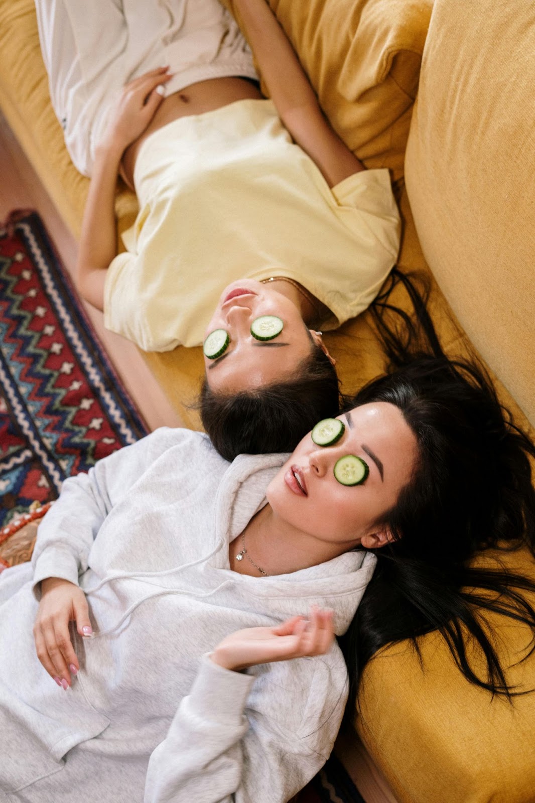 Two young girls lying on the couch relaxing wearing soothing cucumber slices on their eyes.