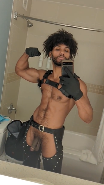 Tony Genius wearing black leather studded chaps and a black gay harness and black gloves while taking a mirror selfie
