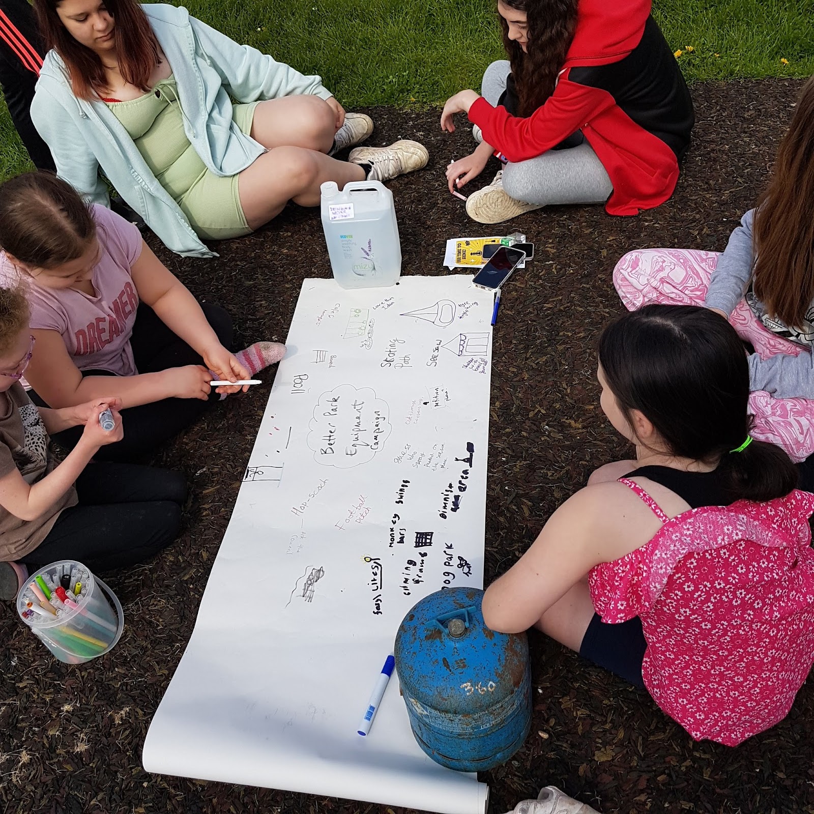 A group of young people sit around a long sheet of paper held in place by a water bottle and a camping gas canister. They are writing and drawing their ideas for new play equipment and other facilities.  
