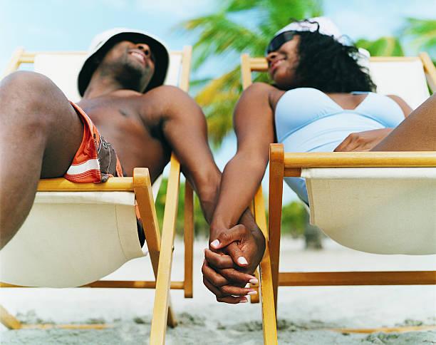couple sitting on deck chairs, holding hands on the beach - black man love stock pictures, royalty-free photos & images