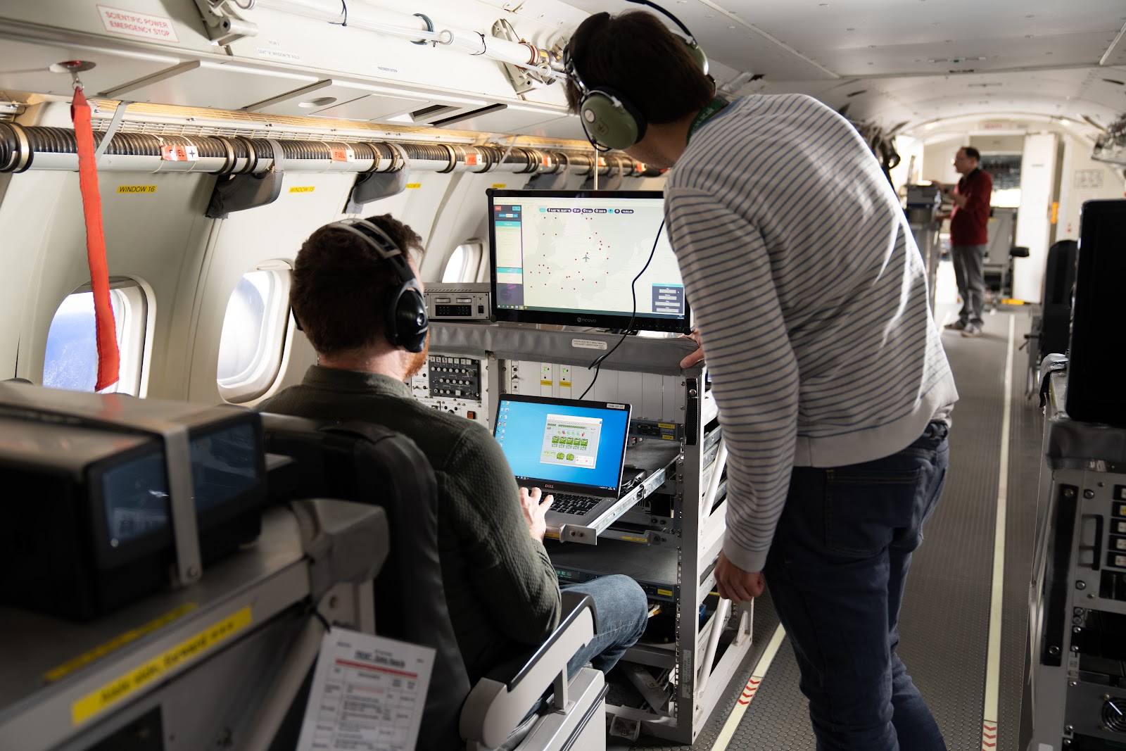 Three people working inside a research aircraft. One person is sat looking at a computer screen with another person standing looking at the screen. One person is standing looking at a laptop screen.