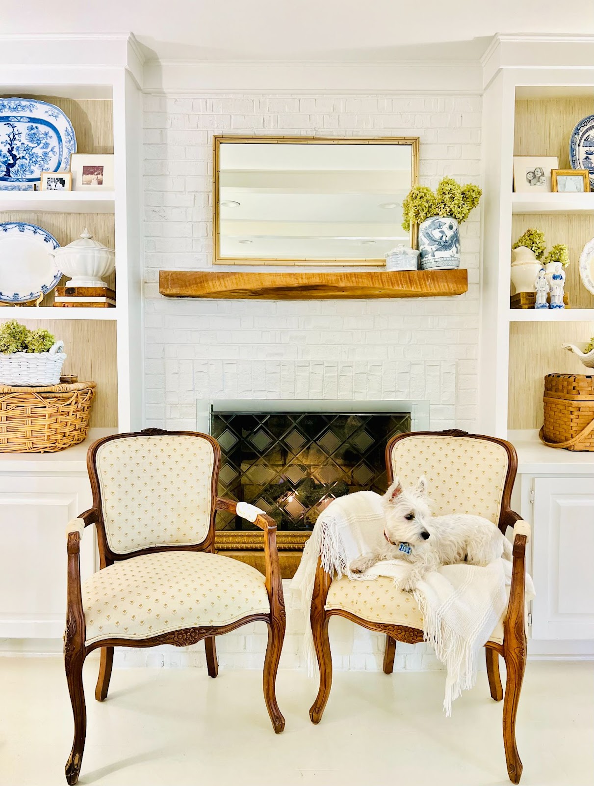 Sally's fireplace adorned with a rectangular gilt mirror, surrounded by an assortment of blue and white porcelains and rattan baskets. Two armchairs are placed in front of the fireplace, with Sally's puppy sitting on one of them.
