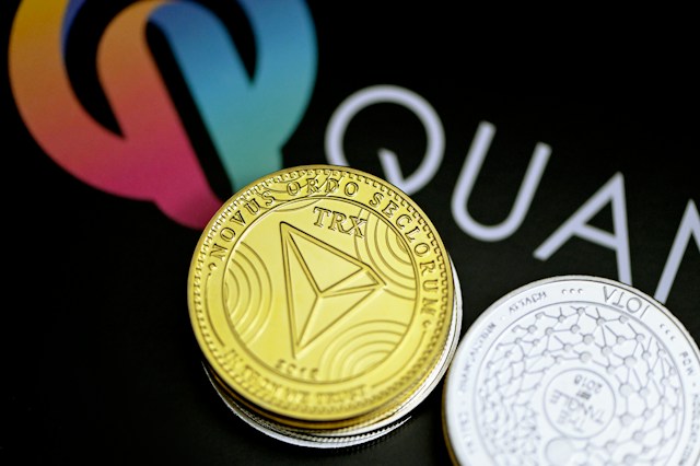 Physical coins representing cryptocurrencies with a Tron coin in the foreground and a Quantum logo in the background.