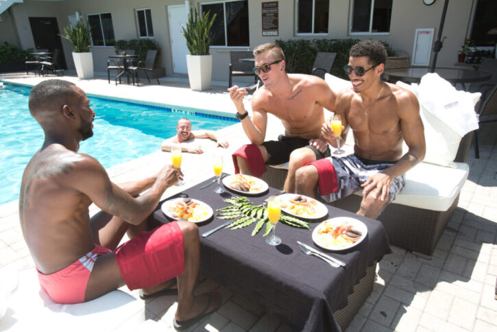 Three gay males wearing swim shorts and speedos by the pool eating breakfast near pool at fort lauderdale clothing optional resort while gay man watches from the pool
