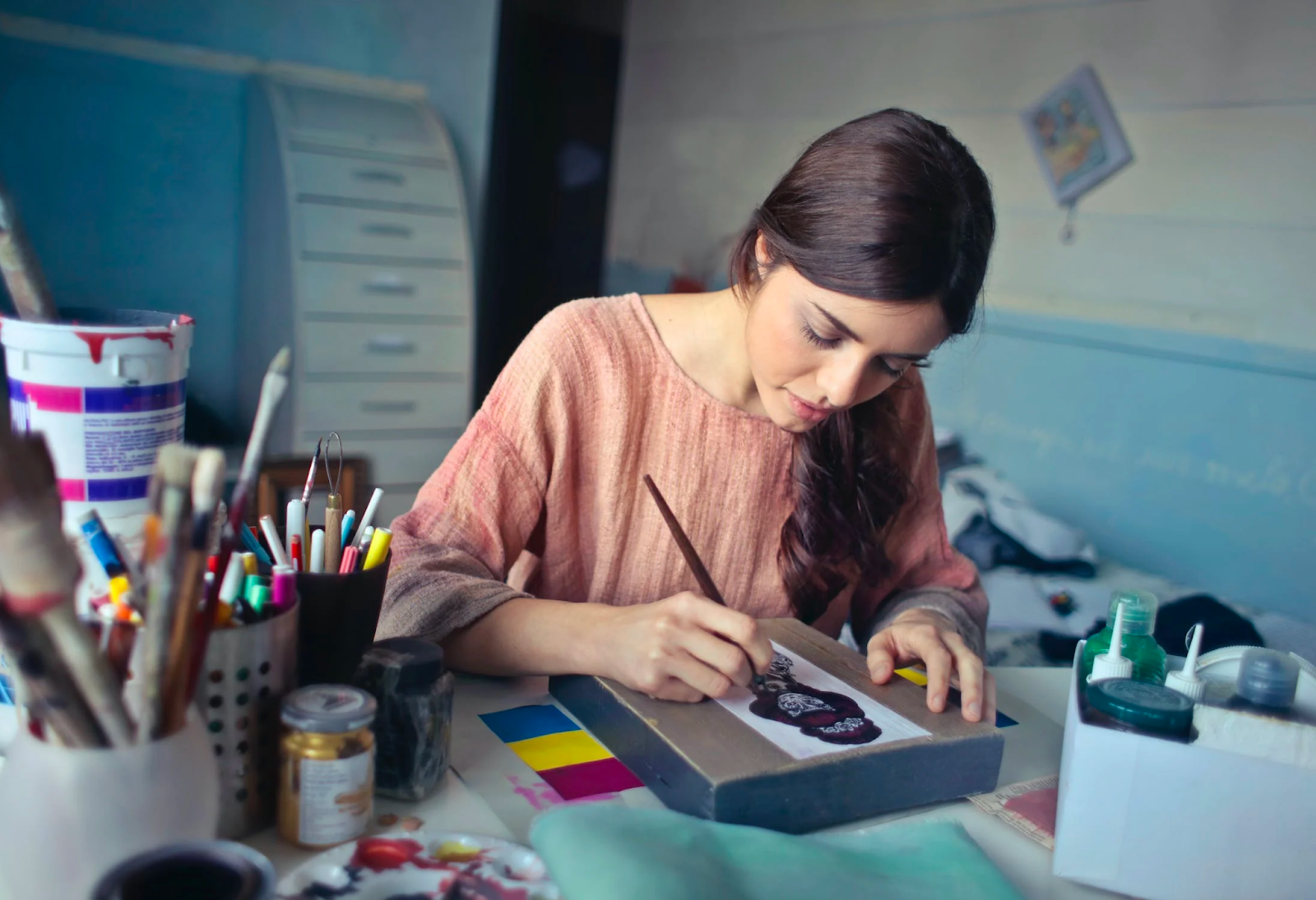 brown haired girl painting at her desk surrounded by brushes