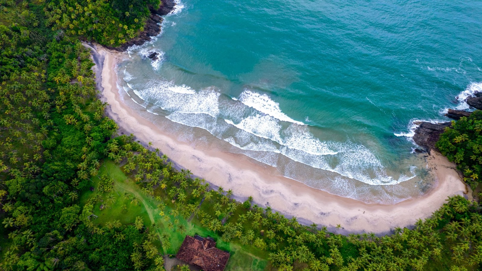 Pequena praia vista de cima, ilustrando o contraste entre a vegetação da Mata Atlântica com o mar azul, faixa de areia branca e ondas que se quebram criando espumas brancas.