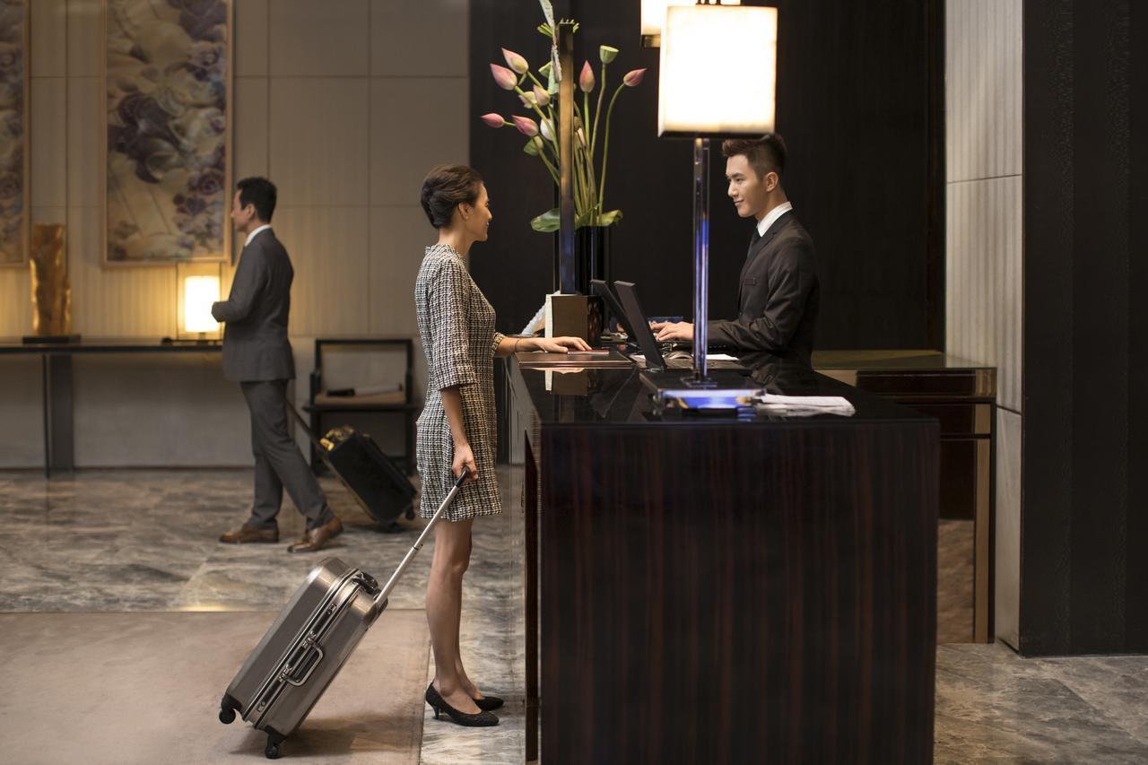 Eine Frau mit kariertem Kleid und Trolley spricht mit dem Angestellten einer Hotelrezeption. Im Hintergrund ein weiterer Mann mit Businessanzug und Trolley. 