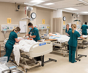 A medical simulation lab with students in lab coats attending to a patient simulator on a hospital bed. Medical equipment is visible in the background.