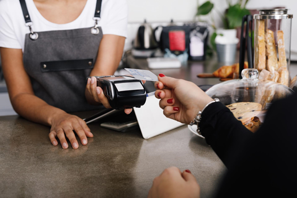 Eine weibliche Hand mit rot lackierten Fingernägeln hält eine American-Express-Kreditkarte an ein Lesegerät. Dieses wird ebenfalls von einer weiblichen Hand gehalten. Die Frauen stehen an einem Verkaufstresen, im Hintergrund sieht man Gebäck bzw. Kaffeekannen. 