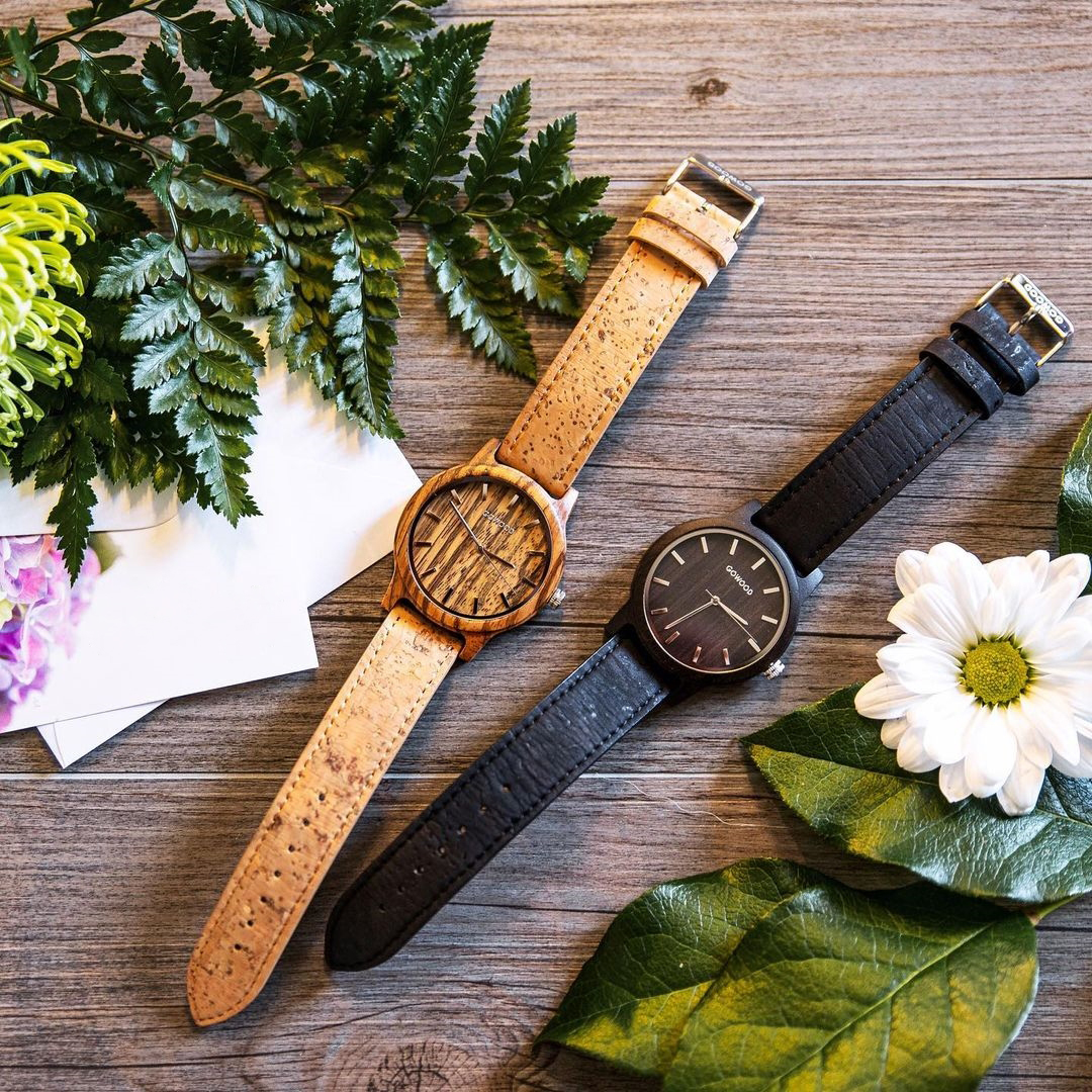 Two gowood watches, one with a light wood face and the other with a dark wood face lie on a table next to a bouquet