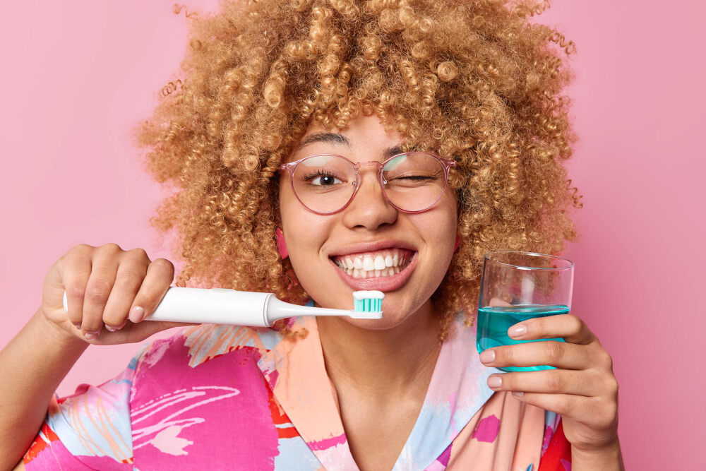 mulher preta com cabelo claro com copo com enxaguante bucal em uma mão e escova de dentes na outra