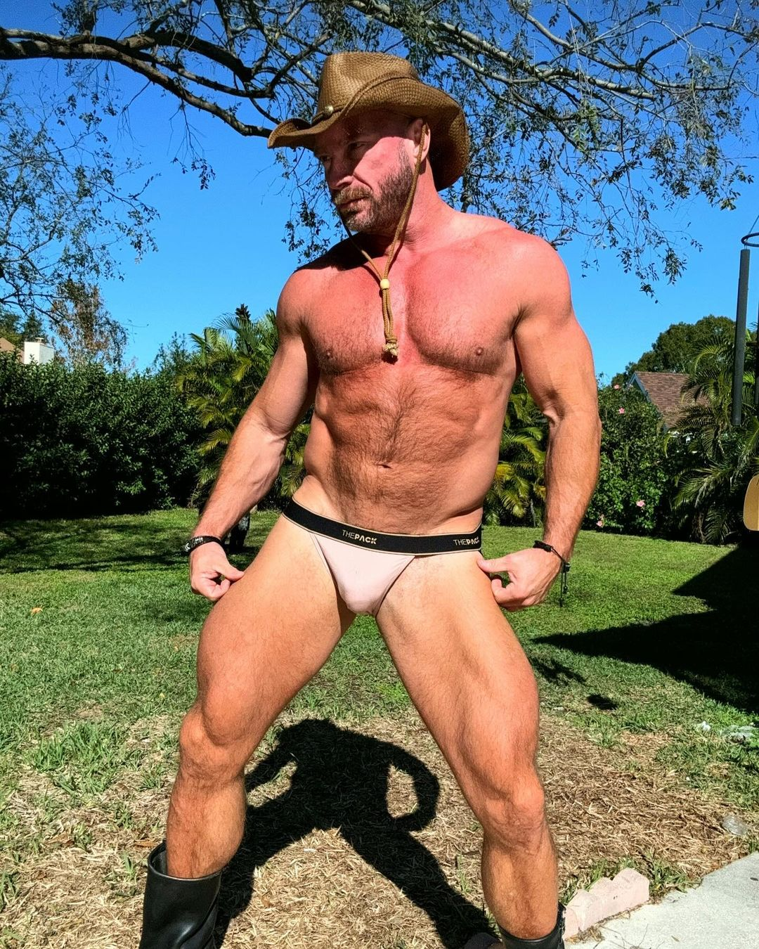 Killian Knox wearing a cowboy hat and cowboy boots in a thepack jockstrap looking into the distance on green grass