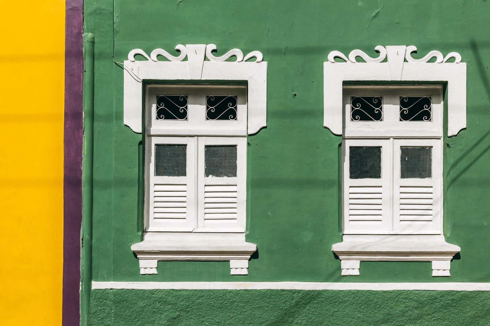 Duas janelas brancas em estilo colonial, em contraste com o verde vibrante da parede. Ao lado esquerdo, uma faixa amarela da construção ao lado.