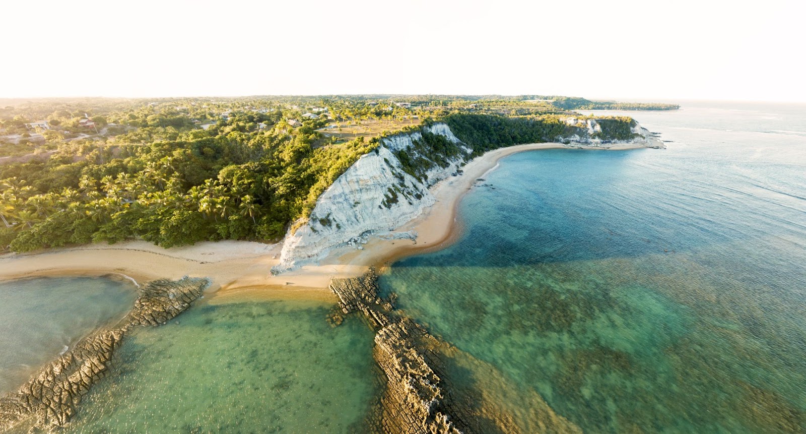 Vista panorâmica de duas praias da Bahia. A maré baixa revela as formações rochosas e corais debaixo do mar esverdeado. Pontal com faixa de areia que contorna a grande falésia.