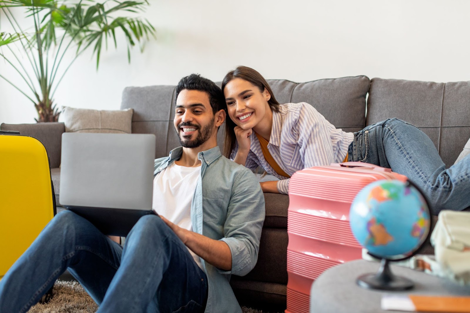 Casal com o homem sentado no chão e apoiado no sofá digitando no notebook apoiado em seus joelhos, com a mulher deitada no sofá também olhando para a tela do notebook. Ao seu redor, malas de viagem e um pequeno globo.