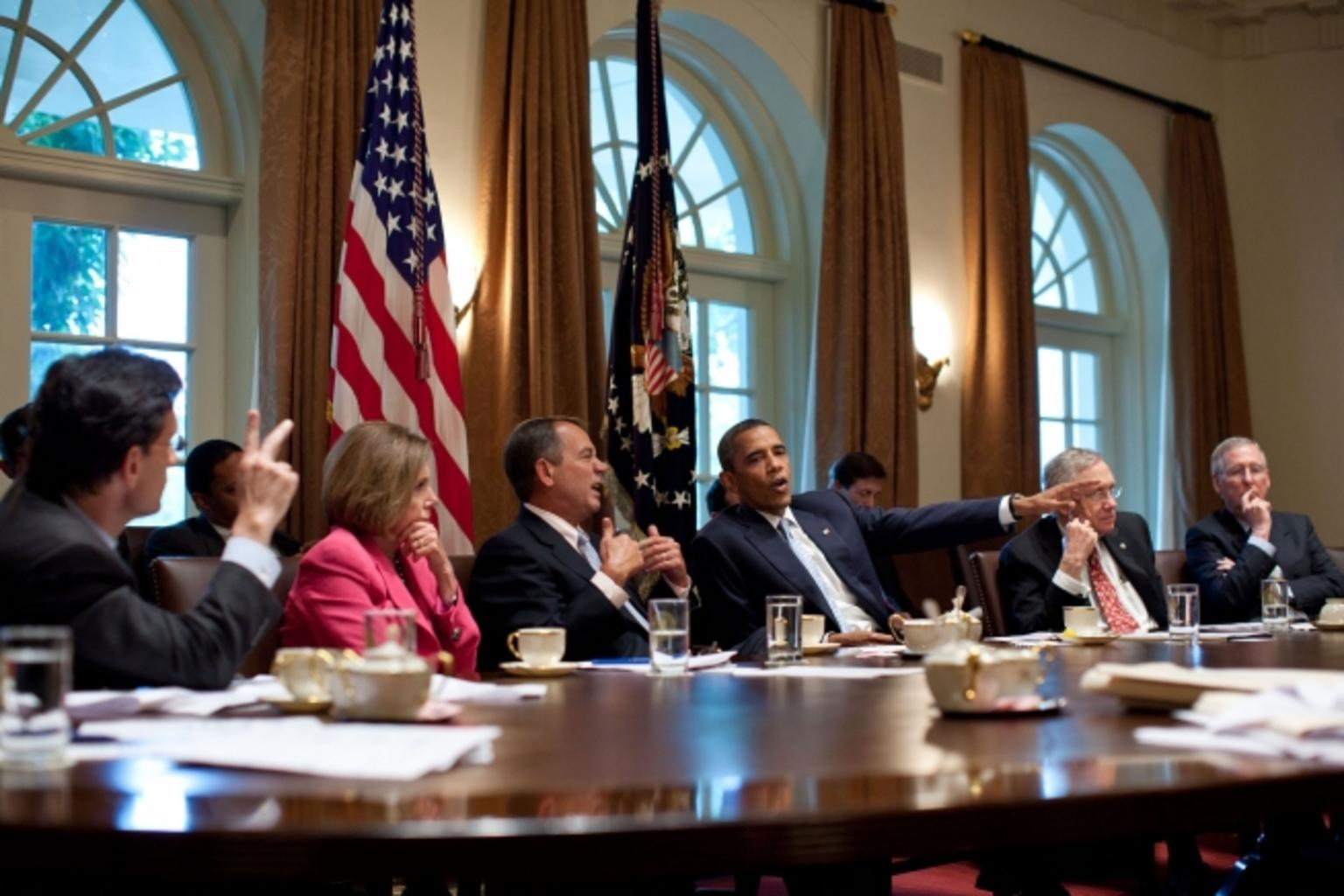 President Obama meets with members of congressional leadership at a large polished wood table. The men are in dark suits and Nancy Pelosi is in bright pink. They have coffee and water in front of them. They appear to be in the middle of a heated discussion. 