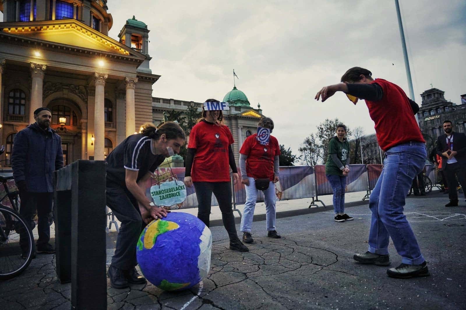 Rebels wear masks showing corporate logos and play football beside parliament. The football is a small planet earth.