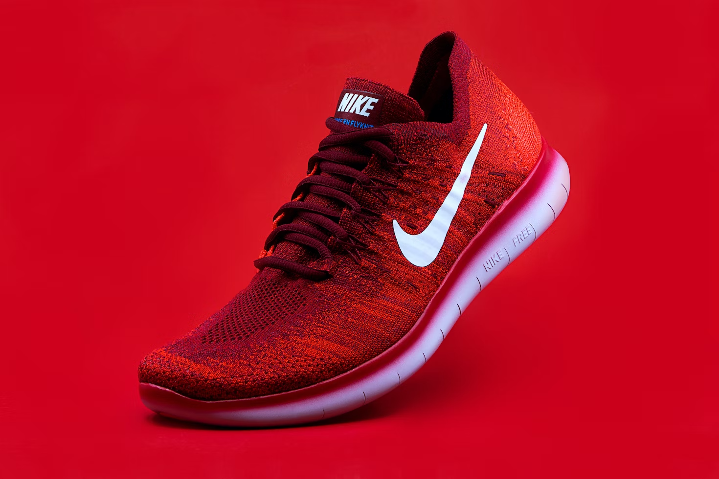 A red Nike Free running shoe against a red background. The shoe has a low-top design with a knit upper that appears to offer breathability and flexibility.