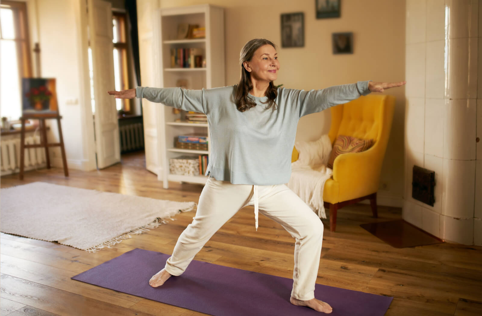 An older adult woman practicing her warrior 2 pose in her living room.