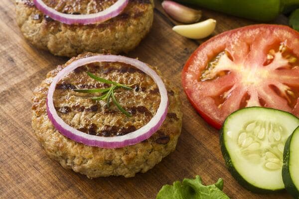 A ground beef patty topped with an onion slice, with sliced cucumbers and tomatoes on the side