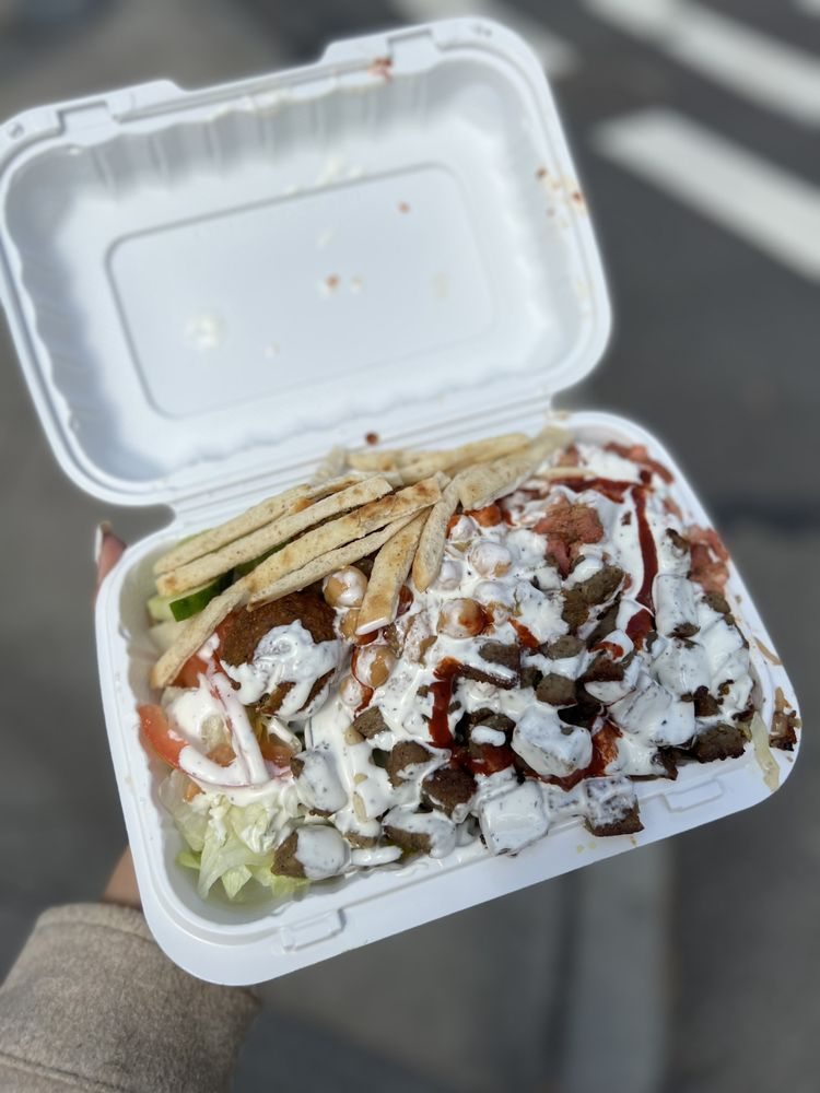 Photo from The Casbah's Yelp page. A clamshell to-go container, open and being held up by a nicely manicured hand. The food looks like pita, chickpeas, falafel, meat, lettuce, cucumber, tomato, covered in sauce.