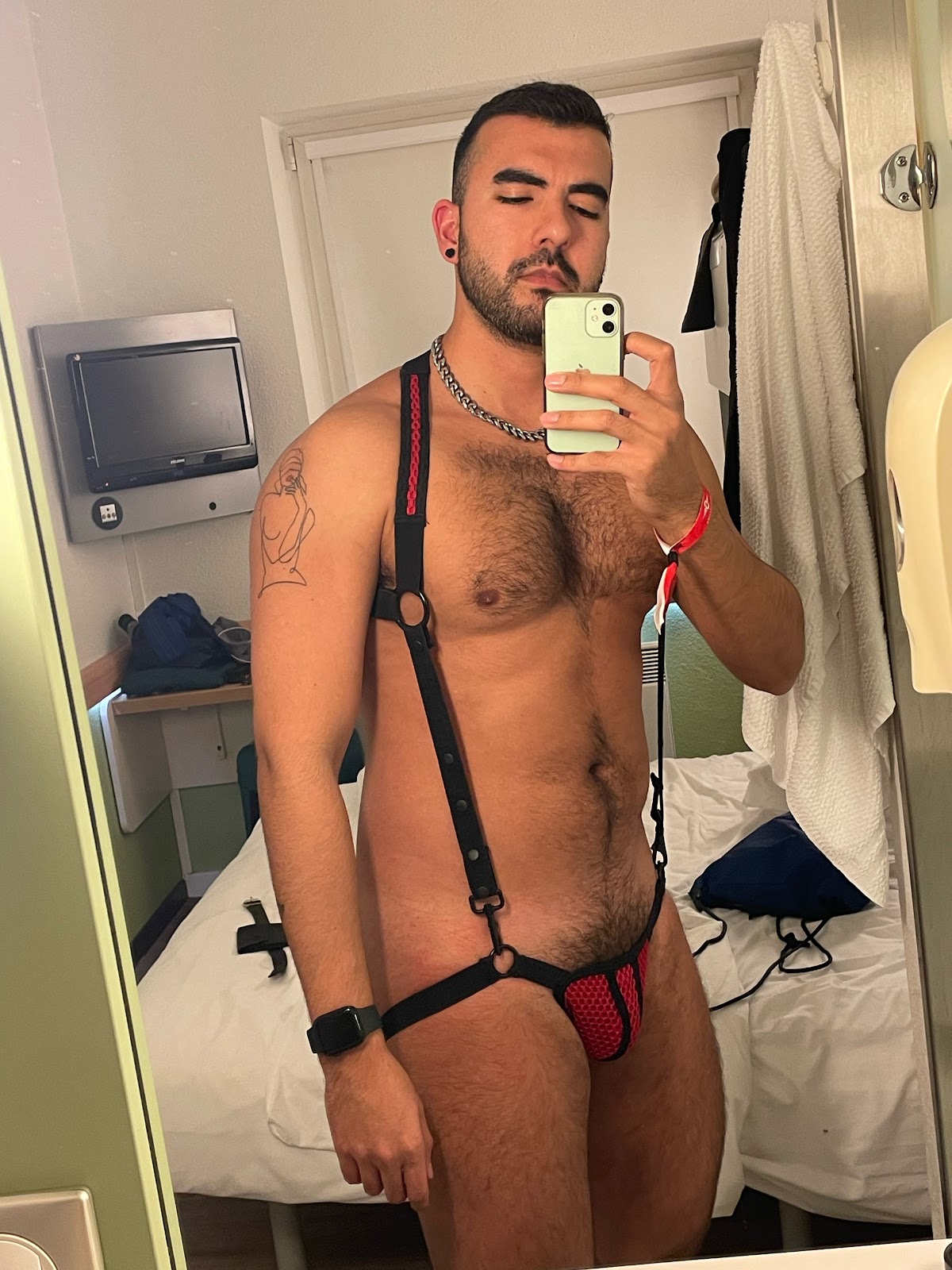 Onlyfans content creator Phil showing off his hairy chest and bush in red and black fetish harness with suspenders taking a selfie in his Belgium hotel room