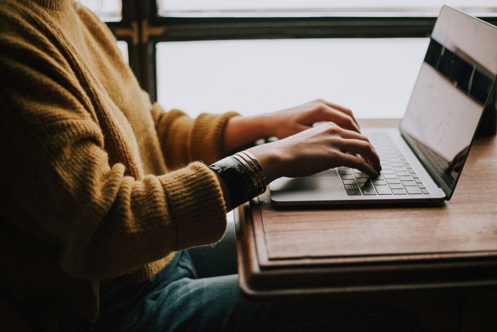  A imagem mostra uma pessoa sentada em uma mesa de madeira, usando um laptop. A pessoa está vestindo um suéter amarelo e calças jeans, com uma mão no trackpad do laptop e a outra mão digitando ou descansando sobre o teclado. Há uma pulseira no pulso da mão que está no teclado. A tela do laptop está visível, mas não é possível discernir o conteúdo específico. O fundo apresenta uma janela com luz natural, sugerindo um ambiente interno com luz do dia.