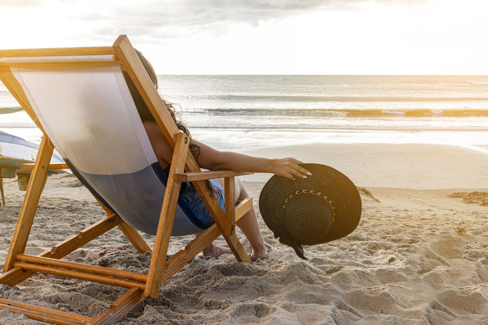 Mulher sentada em cadeira de praia de frente para o mar, segurando seu chapéu na mão.