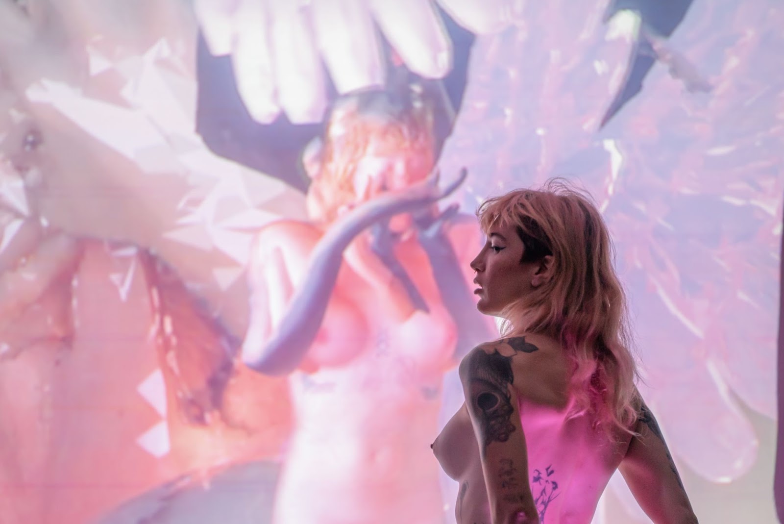 Image: Ava Wanbli performs “Sertraline Dolls'' at Elastic Arts for the Elastro A/V Festival. A 3-D model of a nude trans girl with white wings is projected onto the wall. Ava is standing half-profile and nude with her back to the camera. Her back is illuminated by a pink light. Photo by Mikey Mosher.
