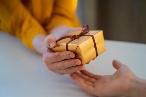 Female hands hold and give a nice packed gift. Female hands hold a gift closeup, top view. A young girl gives a nice packed surprise to her mother, grandmother with love, remembers elderly relatives, visits during holidays, takes care of them. Family Gift stock pictures, royalty-free photos & images