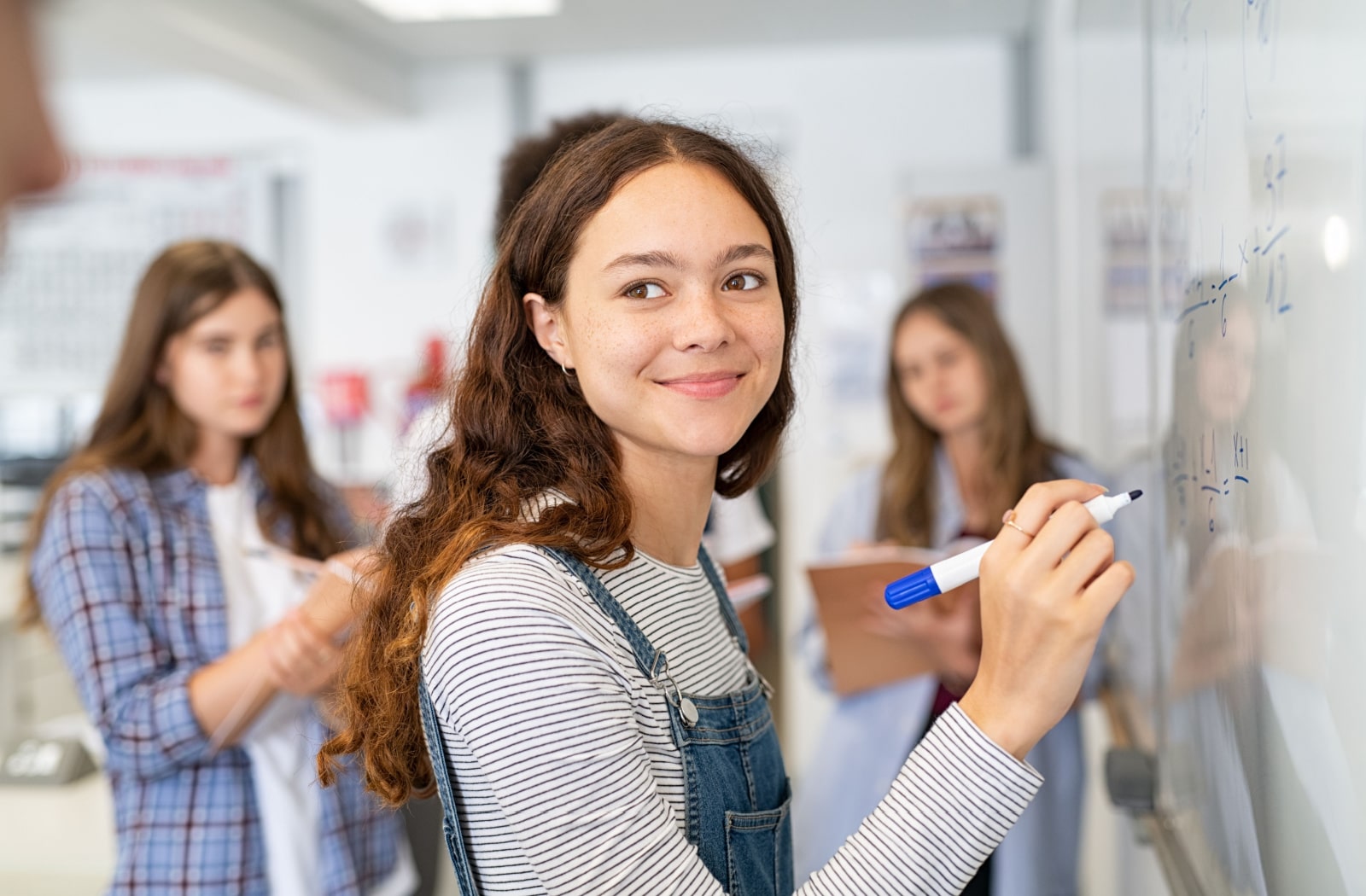 A teenage girl smiling back at someone while she is writing on a dry erase board in a class room