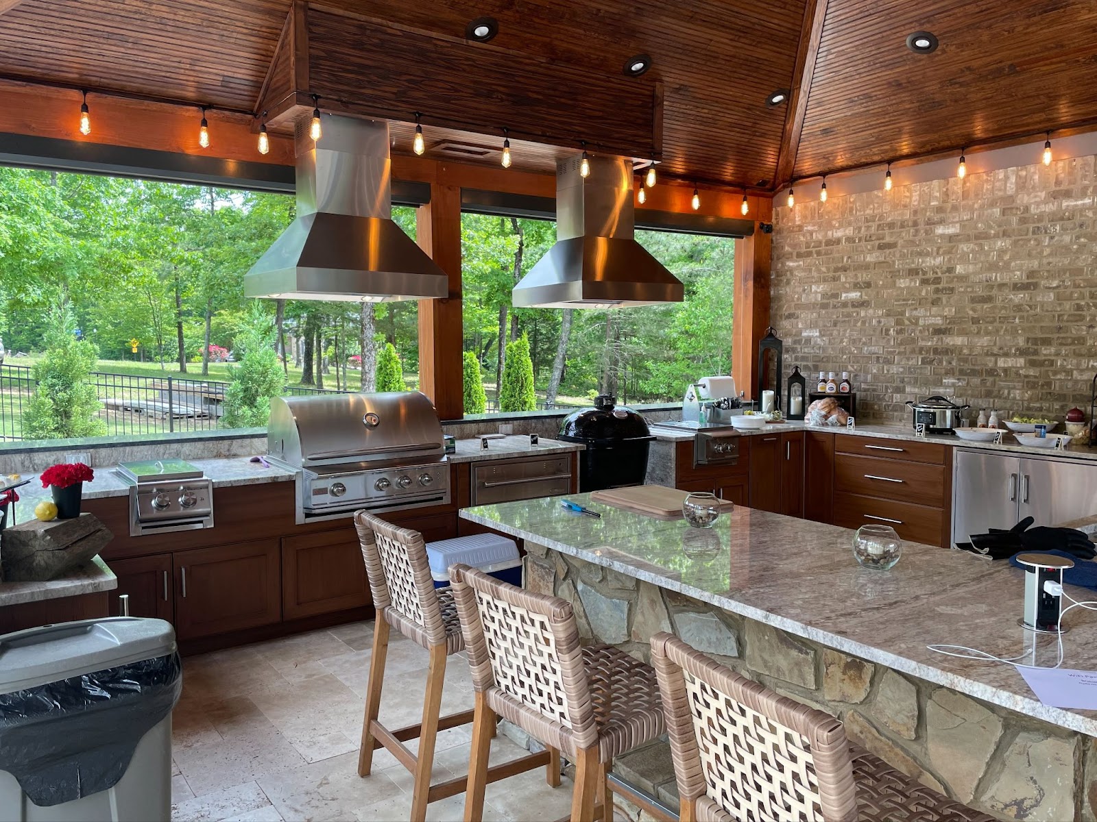 Welcoming open-air cooking space with a natural stone countertop, twin ventilation hoods, and a view of a lush backyard.