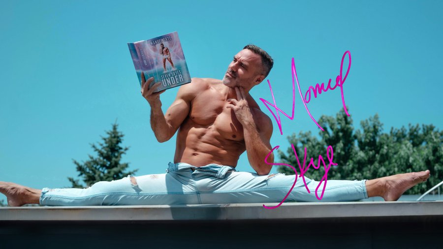 A signed autograph photo of Manuel Skye wearing light denim jeans with the zipper opened reading his book shirtless while sitting in the splits outside