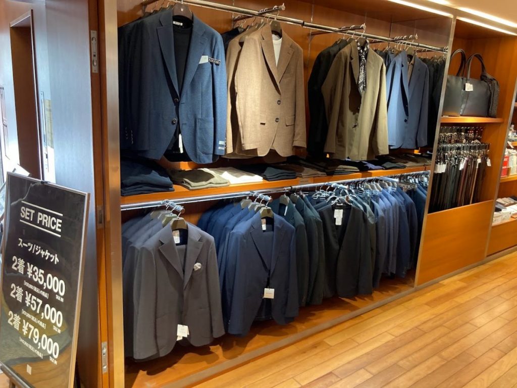 大阪駅・梅田のスーツショップの店内画像。手前にはお得なセットプライスのPOPを設置。奥ではビジネスカジュアルでも着られるスーツをご用意しています。