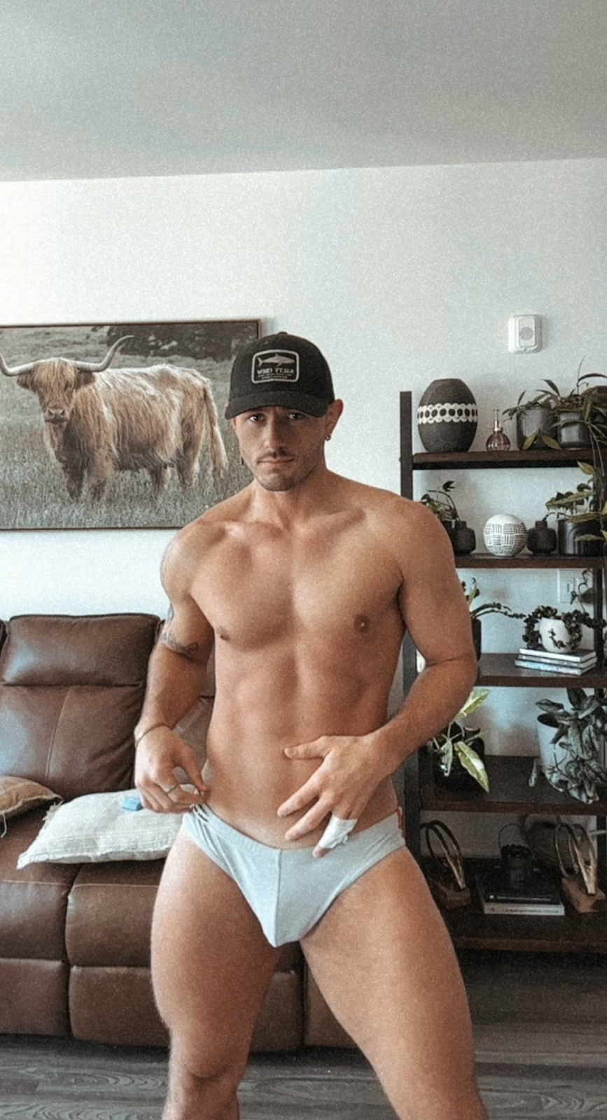 Ryder Owens standing in his grey briefs in his living room wearing a fishing baseball hat and smirking for his onlyfans followers