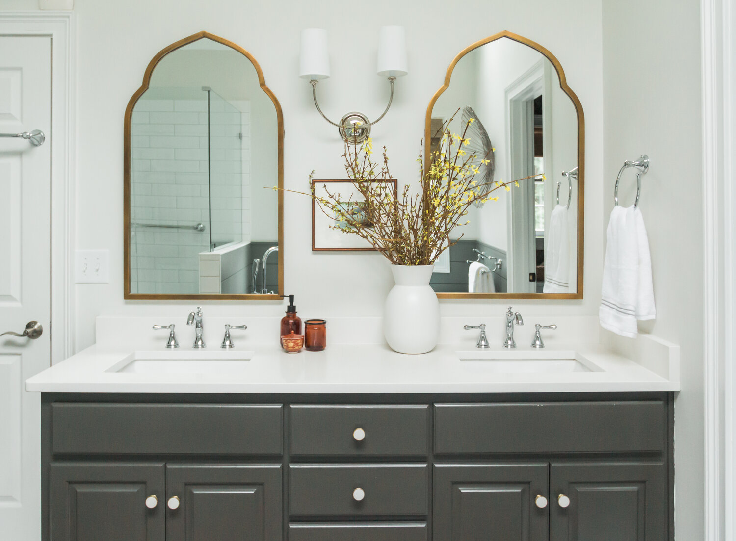 Bathroom vanity with white countertop, dark gray drawers, vintage mirrors, and lampshade lighting. 