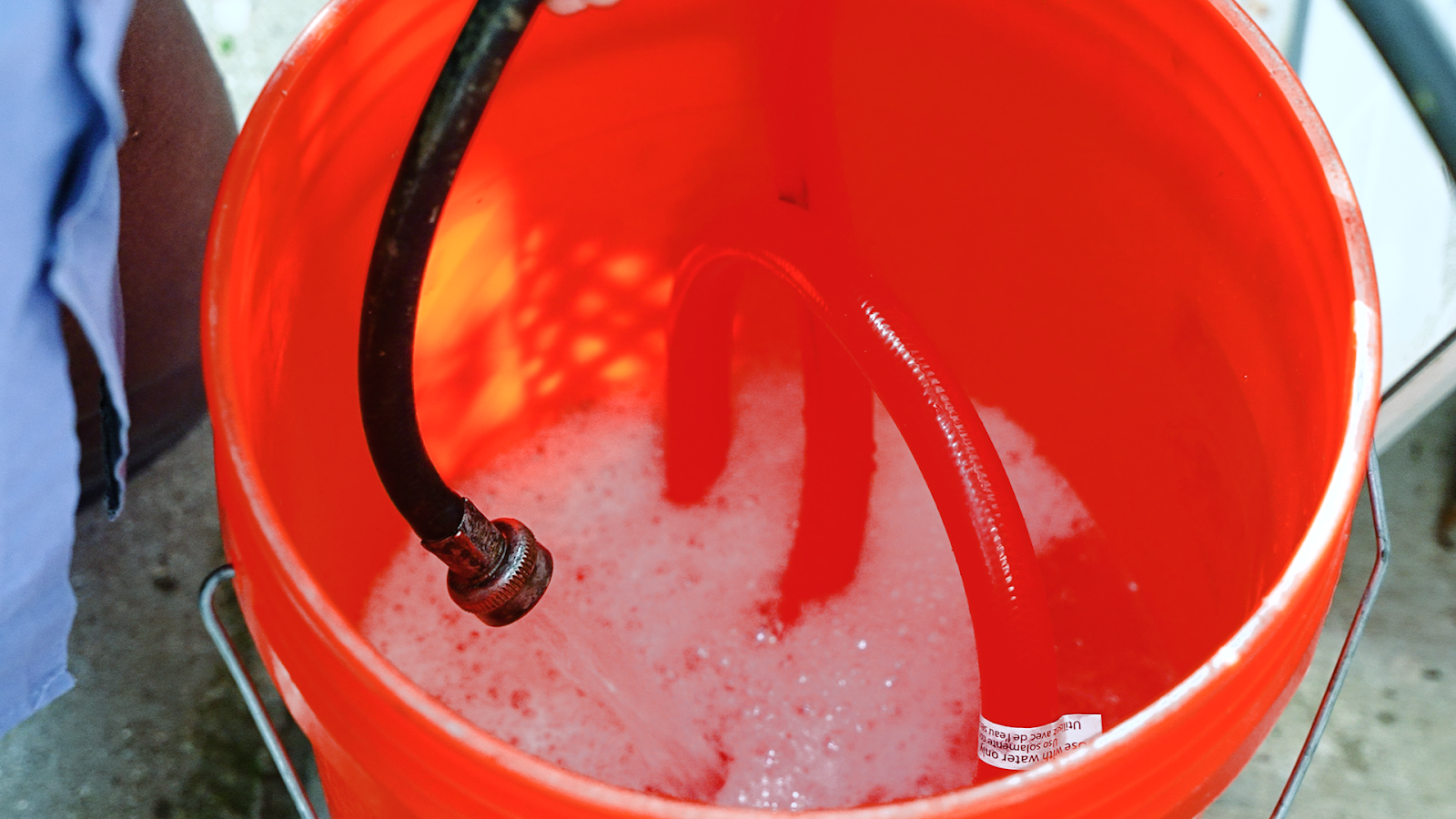 The image shows a black hose draining water from the tankless water heater into a 5-gallon bucket. 