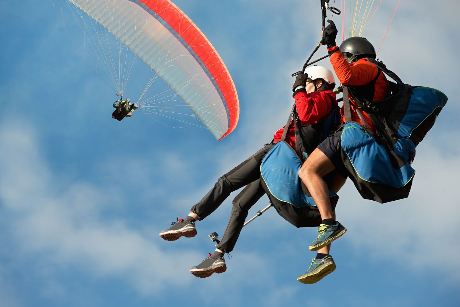 Duas pessoas em foco no ar sobrevoando de parapente. Ao fundo, uma terceira pessoa voando de parapente no céu azul com nuvens brancas.