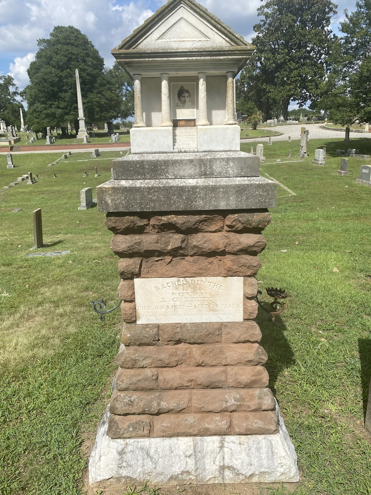 A grave marker with a rough cut marble base and rough cut brown brick.  It is topped with a small replica of a temple and showcases a portrait of Rachael.  It has a name placard that reads Rachael Blythe, wife of A. G. Bauer died January 9, 1897 - aged 26 years.  True worth is being, not seeming.