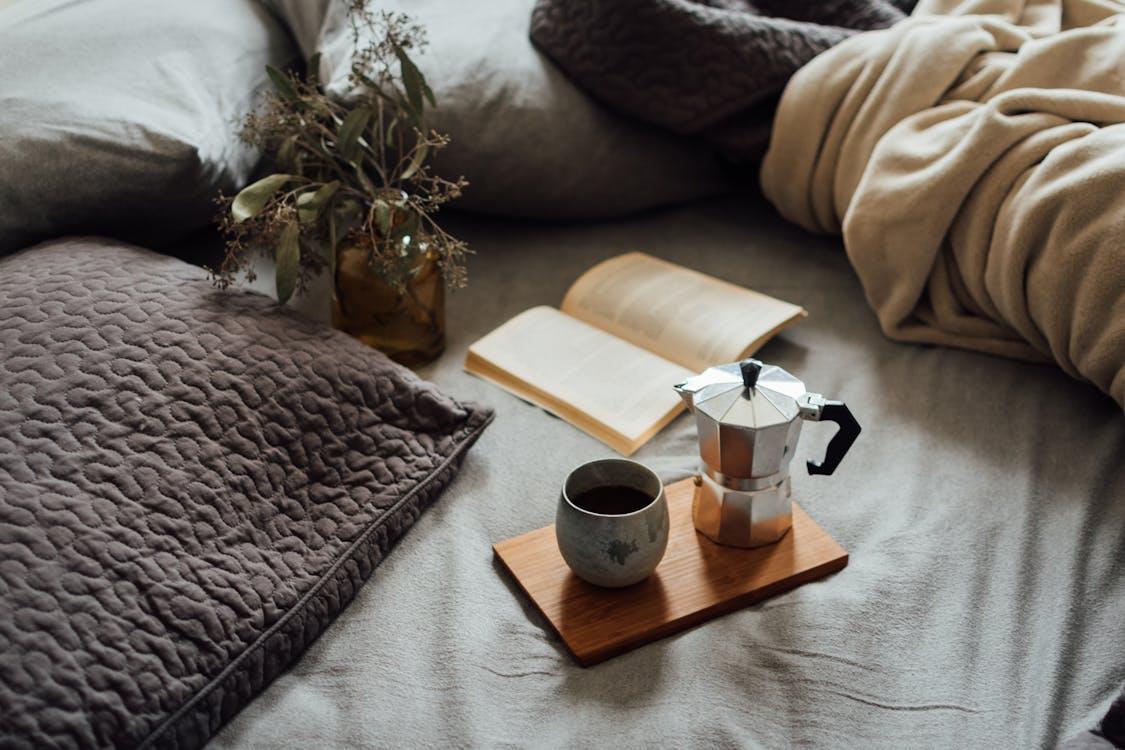 Free From above of cup of hot coffee with pot on wooden tray near open book and dry plant in cozy bed Stock Photo