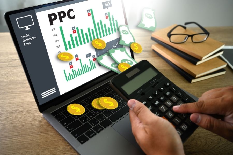 Person using calculator in front of a laptop displaying PPC (Pay-Per-Click) advertising data with graphs, dollar signs, and money icons.