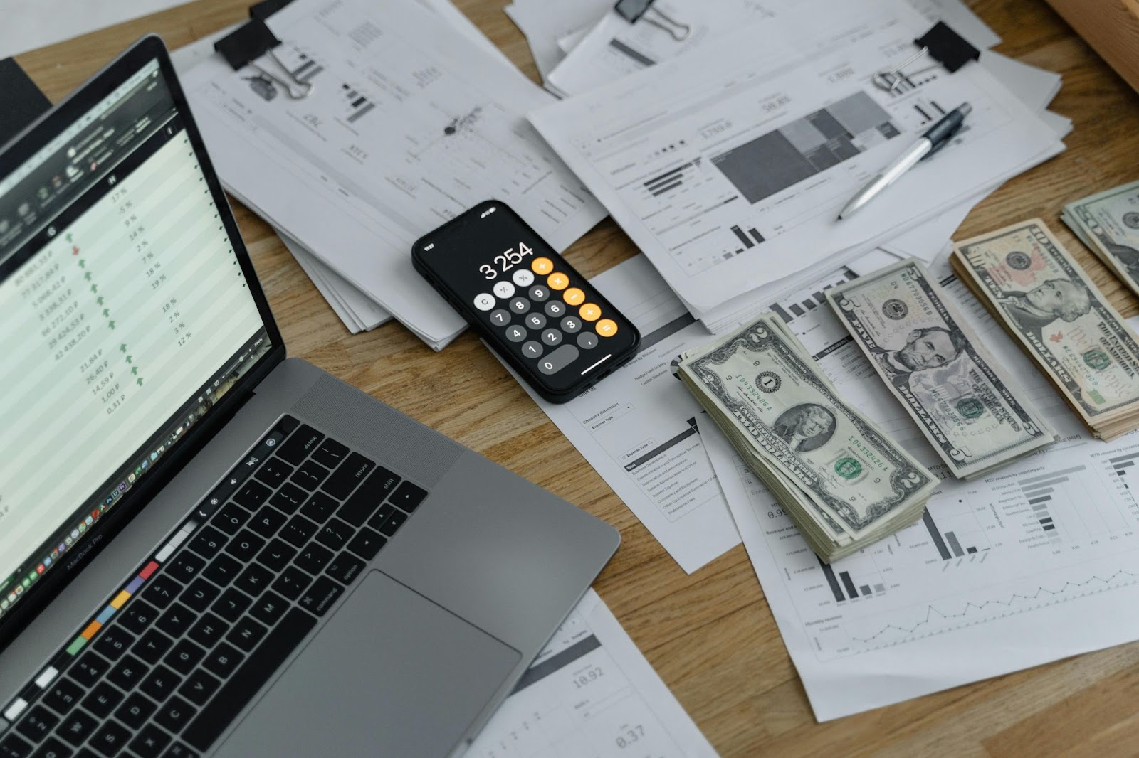 A desktop setup with financial documents, a laptop, a smartphone calculator, and separate stacks of money.