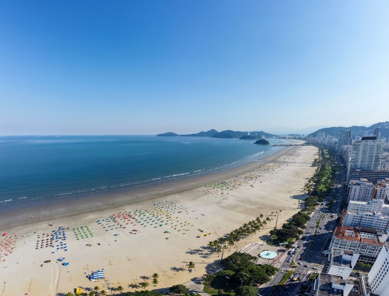 Vista aérea panorâmica da Praia do Gonzaga. Diversas cadeiras de praia de diferentes cores na extensa faixa de areia em frente ao mar azul.