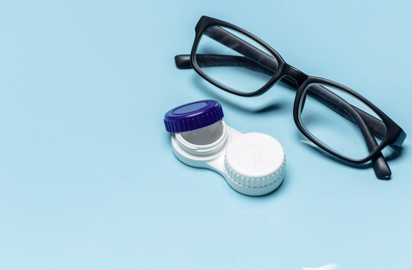 A pair of eyeglasses beside a partially opened contact lens case against a blue background.