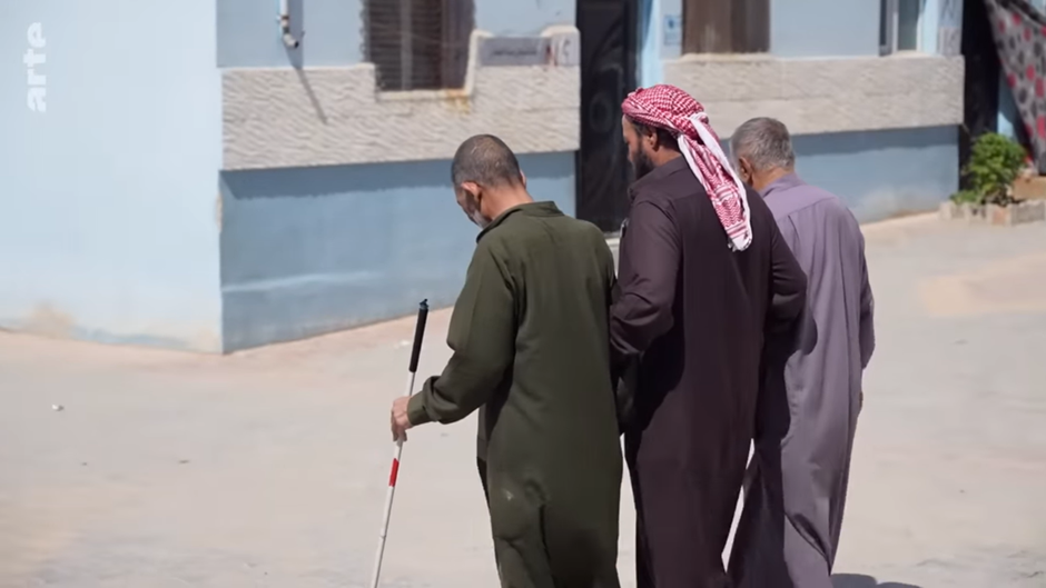 Three men in traditional Middle Eastern robes walking side by side in the streets of Al Nouri refugee camp. The one furthest to the left is holding a mobility cane and the one in the middle is wearing a red keffiyeh (traditional Middle Eastern headdress) around his red.